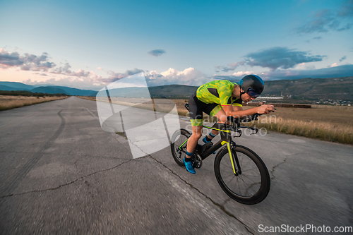 Image of Triathlete riding his bicycle during sunset, preparing for a marathon. The warm colors of the sky provide a beautiful backdrop for his determined and focused effort.