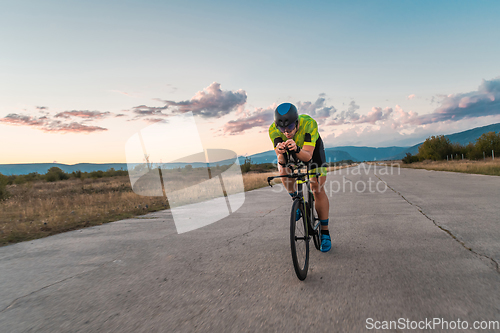 Image of Triathlete riding his bicycle during sunset, preparing for a marathon. The warm colors of the sky provide a beautiful backdrop for his determined and focused effort.