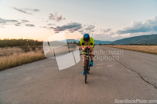 Image of Triathlete riding his bicycle during sunset, preparing for a marathon. The warm colors of the sky provide a beautiful backdrop for his determined and focused effort.