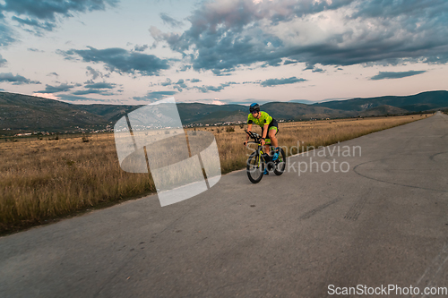 Image of Triathlete riding his bicycle during sunset, preparing for a marathon. The warm colors of the sky provide a beautiful backdrop for his determined and focused effort.