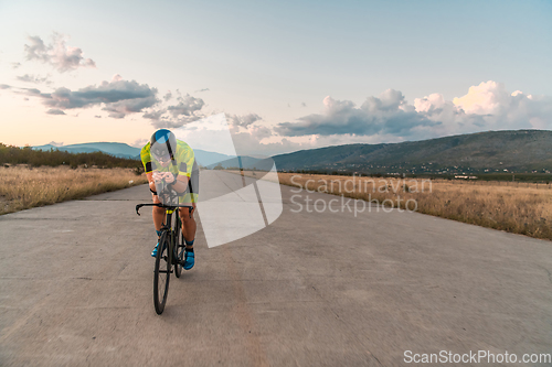 Image of Triathlete riding his bicycle during sunset, preparing for a marathon. The warm colors of the sky provide a beautiful backdrop for his determined and focused effort.