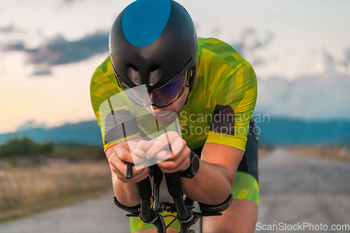 Image of Close up photo of triathlete riding his bicycle during sunset, preparing for a marathon. The warm colors of the sky provide a beautiful backdrop for his determined and focused effort.