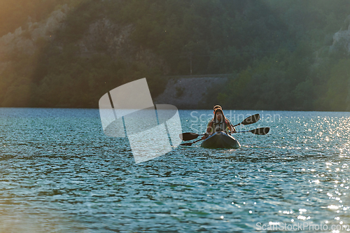 Image of A young couple enjoying an idyllic kayak ride in the middle of a beautiful river surrounded by forest greenery in sunset time