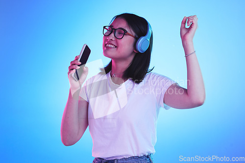 Image of Singing, music and a woman with a phone on a blue background for a podcast, audio or fun. Happy, freedom and an Asian girl with headphones and a mobile app for radio, karaoke or sound on a backdrop