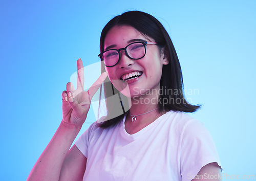 Image of Peace sign, smile and show with portrait of woman in studio for emoji, like and social media. Happy, support and motivation with face of Asian person on blue background for v symbol, gen z and icon