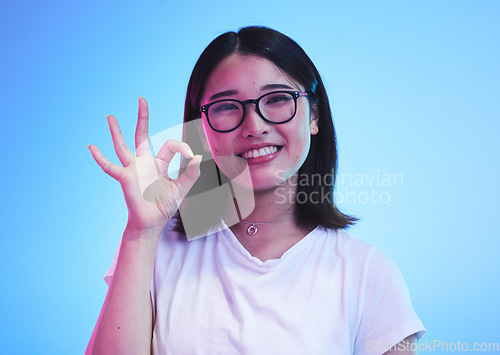 Image of Woman, okay hands or sign for education success, certified and excellence in tshirt and a blue, studio background. Happy young, asian person in glasses, portrait and yes or support emoji for learning