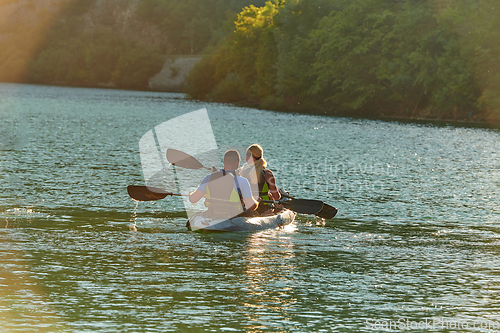 Image of A young couple enjoying an idyllic kayak ride in the middle of a beautiful river surrounded by forest greenery in sunset time