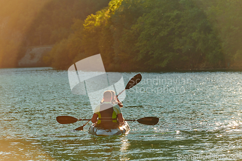 Image of A young couple enjoying an idyllic kayak ride in the middle of a beautiful river surrounded by forest greenery in sunset time