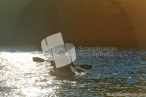 Image of A young couple enjoying an idyllic kayak ride in the middle of a beautiful river surrounded by forest greenery in sunset time