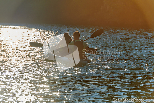 Image of A young couple enjoying an idyllic kayak ride in the middle of a beautiful river surrounded by forest greenery in sunset time