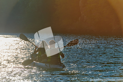 Image of A young couple enjoying an idyllic kayak ride in the middle of a beautiful river surrounded by forest greenery in sunset time