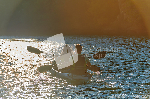 Image of A young couple enjoying an idyllic kayak ride in the middle of a beautiful river surrounded by forest greenery in sunset time