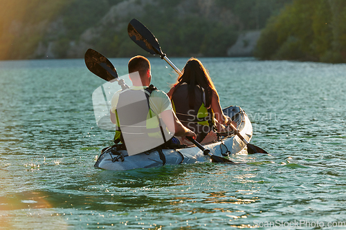 Image of A young couple enjoying an idyllic kayak ride in the middle of a beautiful river surrounded by forest greenery in sunset time