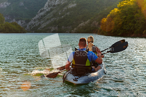 Image of A young couple enjoying an idyllic kayak ride in the middle of a beautiful river surrounded by forest greenery in sunset time