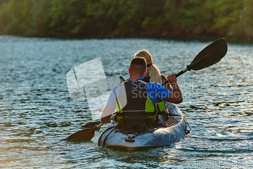 Image of A young couple enjoying an idyllic kayak ride in the middle of a beautiful river surrounded by forest greenery in sunset time