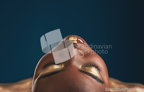 Image of Beauty, gold makeup and art on face of black woman with creative gold aesthetics isolated in a studio blue background. Artistic, cosmetic and young person eyes closed for fantasy with color or paint