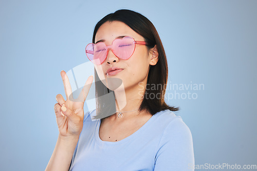 Image of Peace, sign and portrait of woman with hand for emoji in studio blue background with gen z style, fashion or heart glasses. Face, pose and Asian model with cool gesture and expression on social media