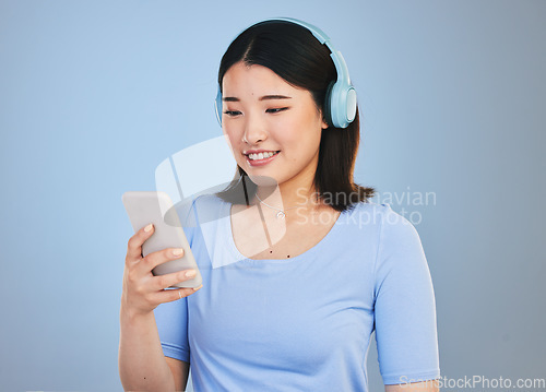 Image of Woman, phone and headphones for listening, audio streaming and social media on a blue background. Young asian person with music subscription, electronics and mobile chat or web networking in studio