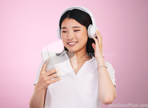 Image of Headphones, smile and woman with a phone in a studio listening to music, playlist or album for entertainment. Happy, calm and Asian female model streaming a song or radio isolated by pink background.