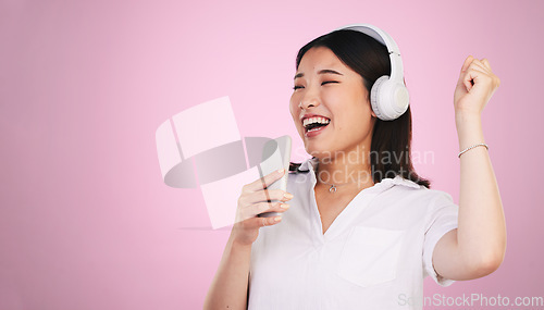 Image of Headphones, singing and woman with a phone in a studio listening to music, playlist or radio. Happy, smile and young Asian female model doing karaoke with a cellphone mic to a song by pink background