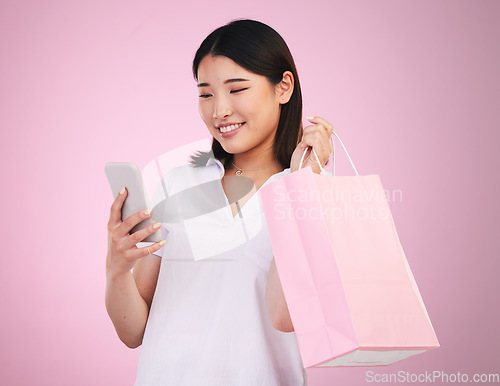 Image of Happy asian woman, phone and shopping bag in payment, purchase or buying against a pink studio background. Female person or shopper on mobile smartphone banking app in ecommerce, sale or discount