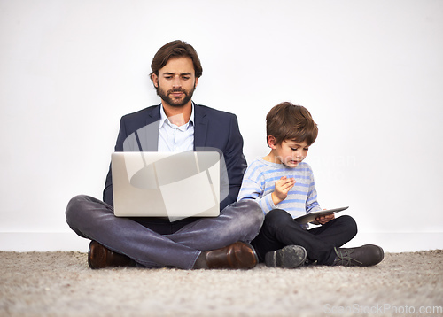 Image of Boys love their digital toys. A father and son sitting on the floor against a wall with a laptop and digital tablet.