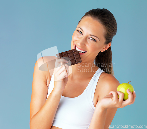 Image of My diet makes room for the occasional indulgence. Studio shot of a fit young woman holding an apple while taking a bite of chocolate against a blue background.