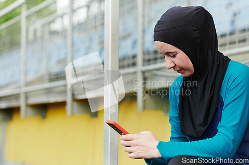 Image of Muslim sports fitness woman dressed in hijab and dark clothes outdoors in green nature park using mobile phone chatting