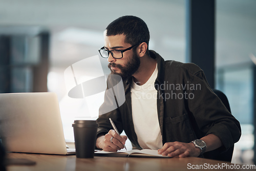 Image of Every success story is created with hard work. Shot of a young businessman writing in a notebook and using a laptop during a late night in a modern office.