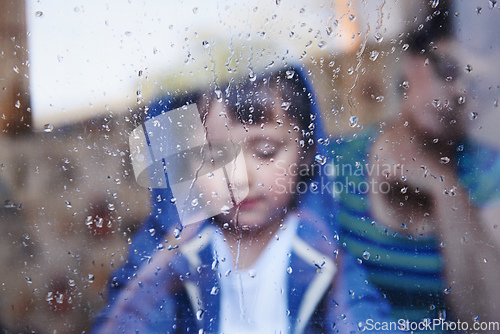 Image of Rainy days with nothing to do.... A little boy looking downwards behind a rain-streaked window while his mother sits in the background.