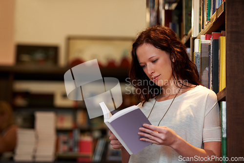 Image of Absorbed in the plot. A young woman lost in the pages of a novel against a backdrop of full bookshelves.