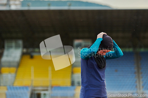 Image of A Muslim woman in a burqa, an Islamic sports outfit, is doing body exercises, stretching her neck, legs and back after a hard training session on the marathon course.