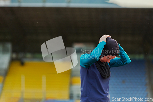 Image of A Muslim woman in a burqa, an Islamic sports outfit, is doing body exercises, stretching her neck, legs and back after a hard training session on the marathon course.