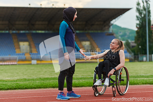 Image of A Muslim woman wearing a burqa supports her friend with disability in a wheelchair as they train together on a marathon course.
