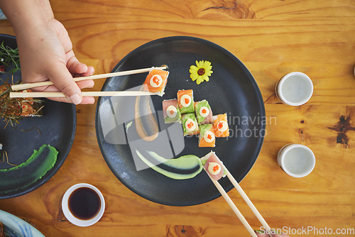 Image of Chopsticks, hand and eating sushi and food at a restaurant for nutrition at table. Above hungry people with wooden sticks and soy sauce for dining, Japanese culture and cuisine with art on a plate