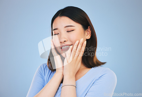 Image of Face, beauty and skincare with an asian woman on a blue background for natural treatment or cosmetics. Smile, makeup and freedom with a young model touching her skin in satisfaction in a studio