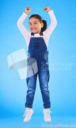 Image of Girl child, jump and celebration in studio with fist in air for goal, achievement or success by blue background. Female kid, excited winner and champion with yes, energy or motivation for competition