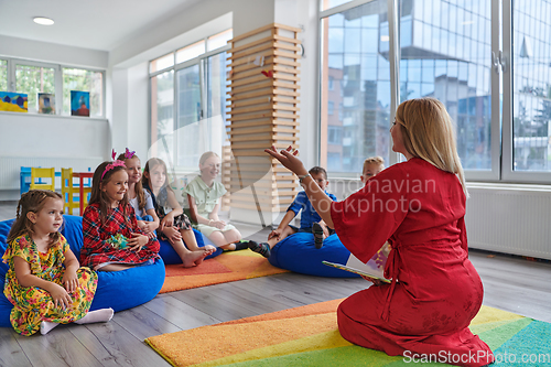 Image of A happy female teacher sitting and playing hand games with a group of little schoolchildren