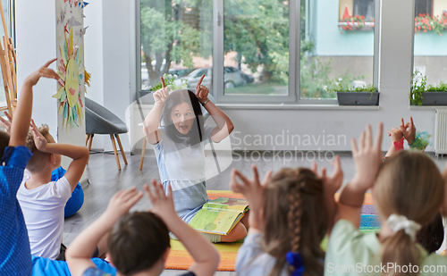 Image of A happy female teacher sitting and playing hand games with a group of little schoolchildren