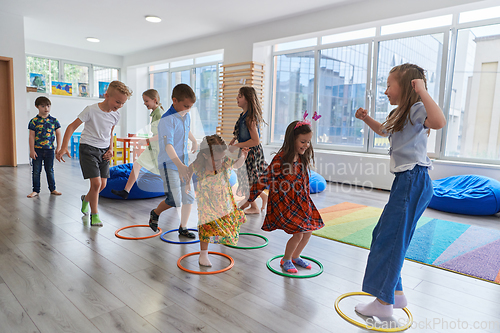 Image of Small nursery school children with female teacher on floor indoors in classroom, doing exercise. Jumping over hula hoop circles track on the floor.