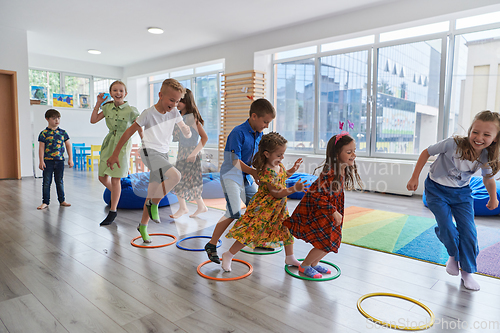 Image of Small nursery school children with female teacher on floor indoors in classroom, doing exercise. Jumping over hula hoop circles track on the floor.