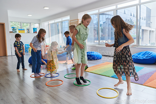 Image of Small nursery school children with female teacher on floor indoors in classroom, doing exercise. Jumping over hula hoop circles track on the floor.