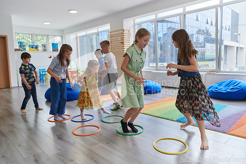 Image of Small nursery school children with female teacher on floor indoors in classroom, doing exercise. Jumping over hula hoop circles track on the floor.