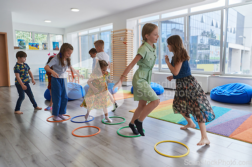 Image of Small nursery school children with female teacher on floor indoors in classroom, doing exercise. Jumping over hula hoop circles track on the floor.