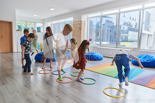 Image of Small nursery school children with female teacher on floor indoors in classroom, doing exercise. Jumping over hula hoop circles track on the floor.