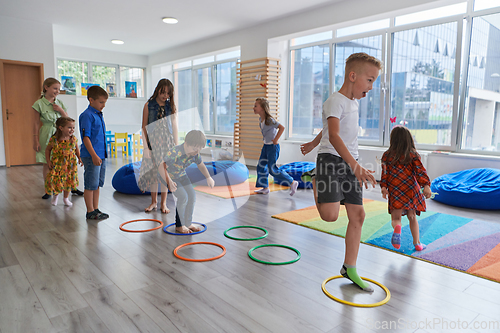 Image of Small nursery school children with female teacher on floor indoors in classroom, doing exercise. Jumping over hula hoop circles track on the floor.