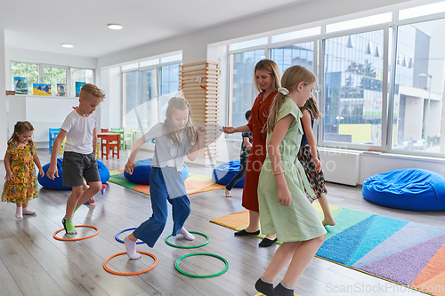 Image of Small nursery school children with female teacher on floor indoors in classroom, doing exercise. Jumping over hula hoop circles track on the floor.