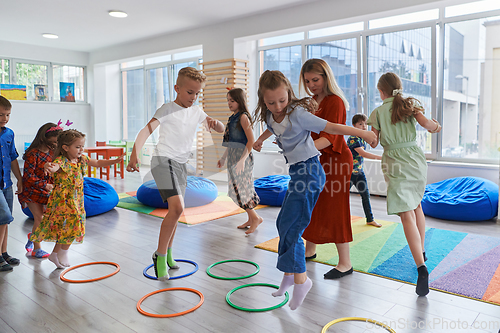 Image of Small nursery school children with female teacher on floor indoors in classroom, doing exercise. Jumping over hula hoop circles track on the floor.