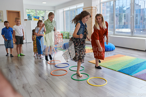 Image of Small nursery school children with female teacher on floor indoors in classroom, doing exercise. Jumping over hula hoop circles track on the floor.