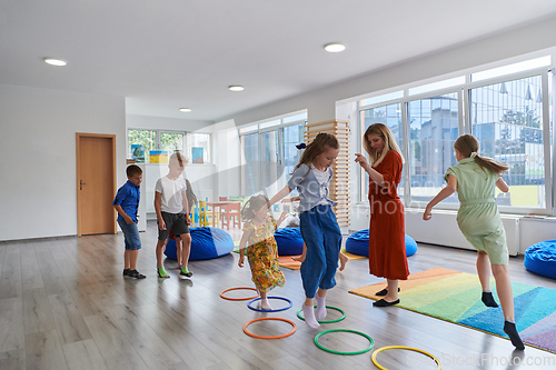 Image of Small nursery school children with female teacher on floor indoors in classroom, doing exercise. Jumping over hula hoop circles track on the floor.